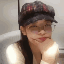 a woman wearing glasses and a hat is sitting with her hand on her chin .