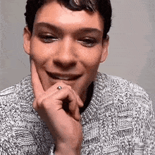 a man wearing a sweater and a ring on his finger is smiling with his hand on his chin .