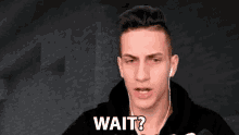 a young man wearing ear buds and a black hoodie is asking someone to wait .