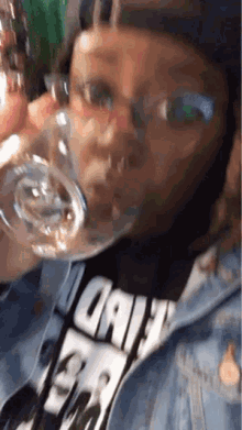 a woman wearing glasses and a denim jacket is drinking from a glass .