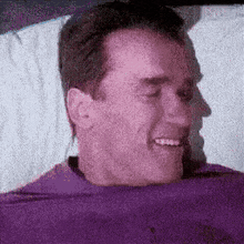 a man is laying in a bed with a purple shirt on and smiling .