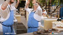 two men in blue aprons are dancing in a kitchen with a nbc logo in the background