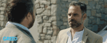a man in a suit talks to another man in front of a stone wall with greed written on the bottom