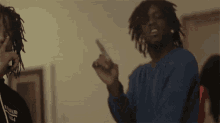two men with dreadlocks are standing next to each other in a room . one of the men is pointing at the camera .