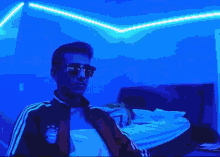 a man wearing sunglasses and a red jacket is sitting on a bed in a bedroom with blue lights .