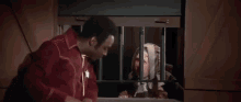 a man in a red jacket is standing next to a woman in a jail cell .