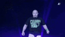 a bald man wearing a pissari not dead t-shirt stands in front of a crowd