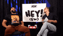 two men sitting in front of a sign that says hey