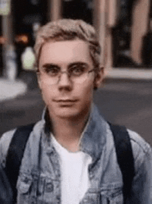 a young man wearing glasses and a denim jacket is standing on the street .