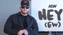a man wearing sunglasses and a hat stands in front of a sign that says hey ( ew )