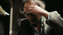 a man wearing glasses holds his hand to his forehead in front of a #kalkazan sign