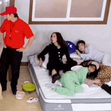a man in a red shirt is standing next to a group of people laying on a mattress