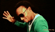 a man wearing sunglasses and a green sweater has the website takasmoke.org at the bottom of his screen