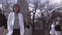 a woman in a white lab coat is standing in a park with a statue in the background .