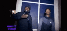 two men are standing next to each other in front of a window in a dark room .