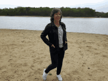 a woman in a black jacket stands on a sandy beach near a lake