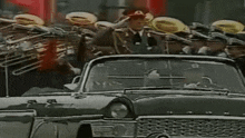 a man in a red hat salutes while driving a car in front of a marching band .
