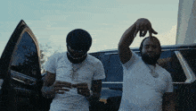 two men are standing next to each other in front of a car and one is wearing a mask