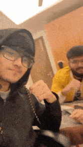 a man wearing glasses and a hoodie is taking a selfie with another man while sitting at a table .