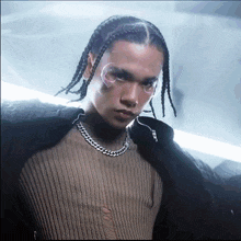 a young man with braids and a chain around his neck looks at the camera