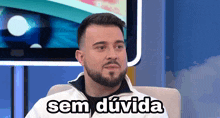 a man with a beard is sitting in front of a television with the words sem duda written on his face .