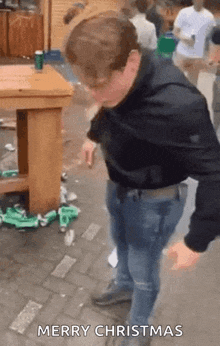 a man is standing in front of a table with bottles on the ground and a sign that says merry christmas .