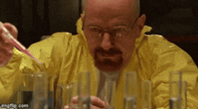 a man in a yellow coverall is pouring liquid into beakers .