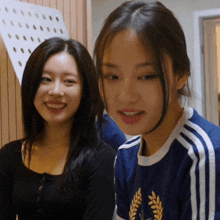 a woman in a blue shirt with a laurel wreath on it smiles next to another woman