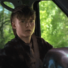 a young man in a plaid shirt is sitting in the driver 's seat of a car looking out the window