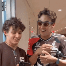 two young men wearing sunglasses are standing next to each other and looking at a cell phone .