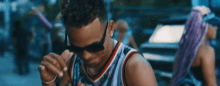 a man wearing sunglasses and a basketball jersey is standing in front of a car .
