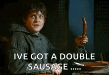 a man is sitting at a table holding a sausage and saying `` ive got a double sausage '' .