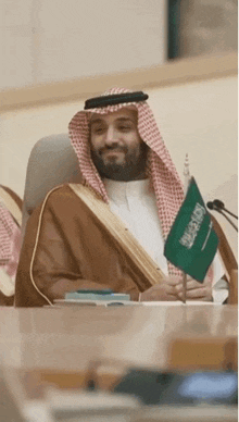 a man in a traditional arabic dress is sitting at a table holding a small flag .