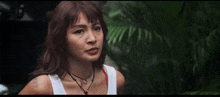 a woman in a white tank top is standing in front of a palm tree and looking at the camera .