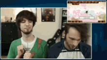 two men are playing a video game and one of them is wearing a green shirt with the letter x on it