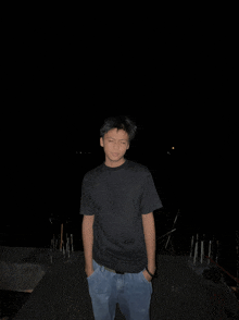 a young man wearing a black shirt and blue jeans stands in the dark with his hands in his pockets