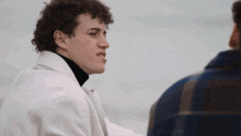 a man in a white coat and a black turtleneck is talking to another man .