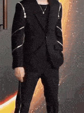 a man wearing a black suit and a necklace is standing in front of a fire background .