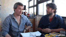 two men are sitting at a table with a book and a glass of beer ..