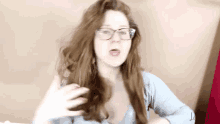 a woman with long red hair and glasses is wearing a gray sweater and making a funny face .