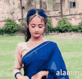 a girl in a blue saree is standing in a grassy field with the word alamin in the corner