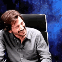 a man in a grey shirt is sitting in a chair and smiling .