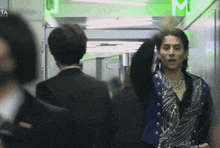 a man in a blue vest stands in a hallway with a green m on the wall behind him