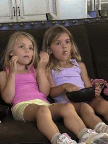 two young girls are sitting on a couch eating popcorn