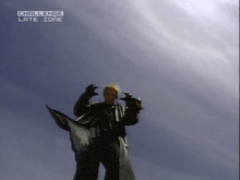 a man in a black coat stands in front of a blue sky with the words challenge late zone above him
