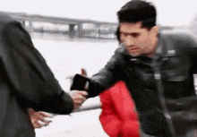 a man in a black jacket is shaking hands with another man in a red jacket while standing next to a body of water .