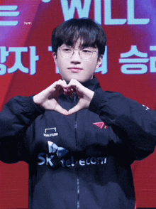 a young man wearing glasses and a sk telecom jacket makes a heart with his hands
