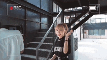 a woman wearing a black shirt that says j.cups stands in front of stairs