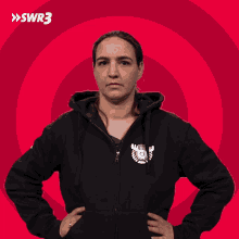 a woman wearing a black hoodie stands with her hands on her hips in front of a red background with swr3