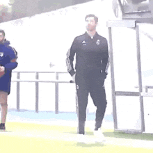 a man in a black adidas tracksuit is standing on a soccer field .